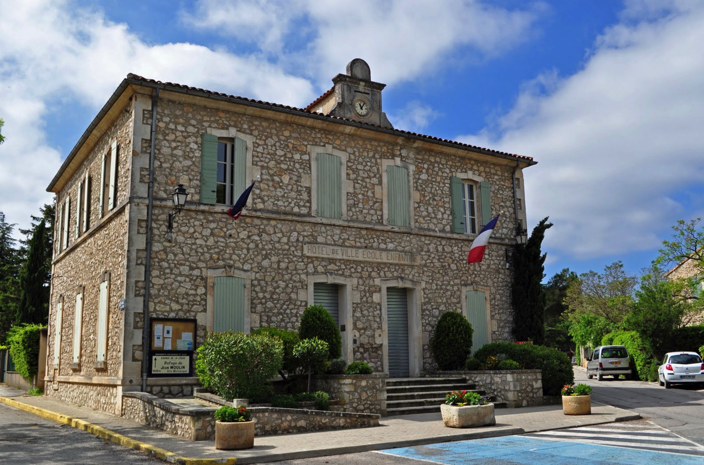 A two-story stone building with a french flag, labeled 'mairie de goult,' flanked by blooming potted plants and a blue sky with scattered clouds.